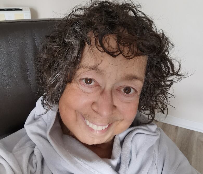 A woman with short, dark and wavy hair smiles at the camera. She is wearing a grey hooded sweatshirt and appears to be sitting on a dark leather chair.