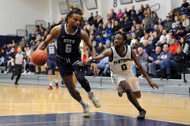 Two basketball players jostle for the ball on a hardwood court. A crowd watches the game on the bleachers behind them. 