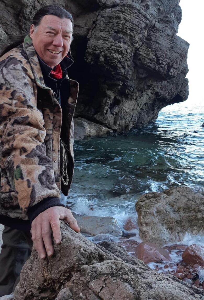 An Indigenous man stands by a rock with a rock cliff behind him.