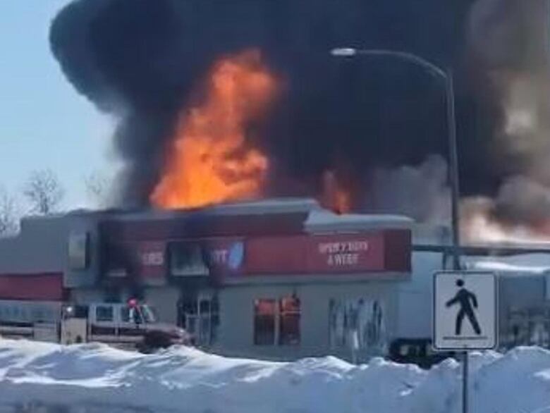 A large orange flame comes out of a Shoppers Drug Mart store in winter as black smoke billows around it.