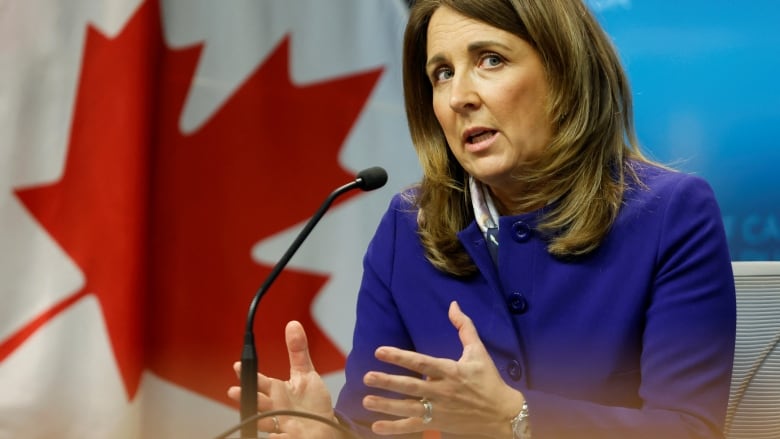 A woman speak at a microphone with a Canadian flag in the background.