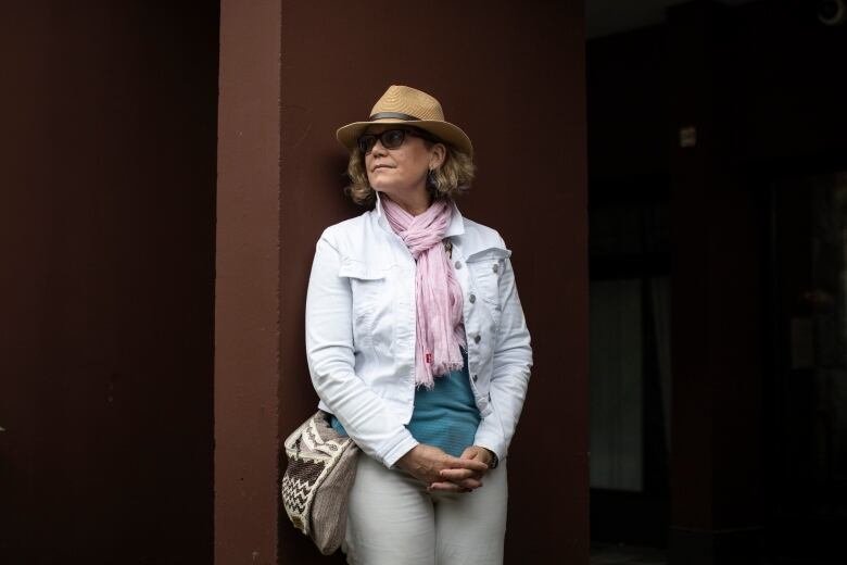 A white woman with chin-length blonde hair is shown leaning against a brown pillar with her hands clasped in front of her. She wears a straw hat, white jeans and white denim jacket, and a pink scarf over a teal shirt.