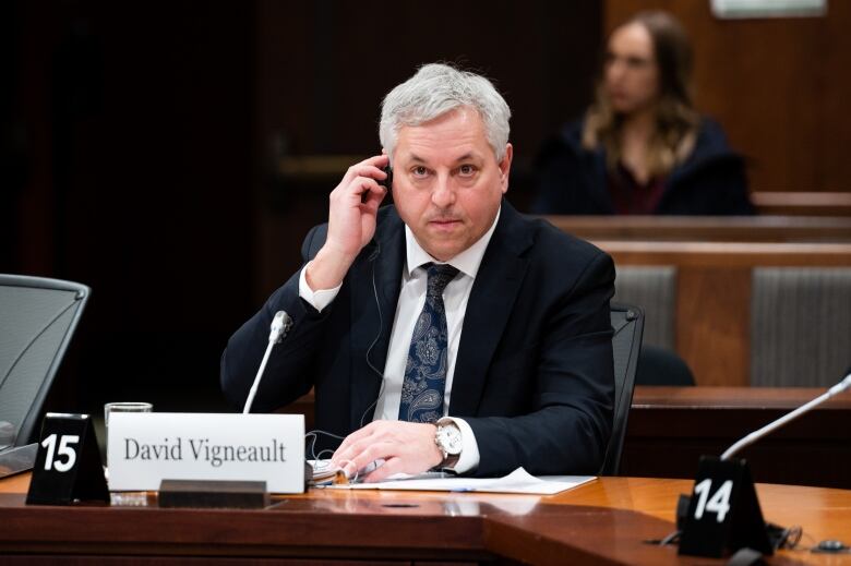 Director of the Canadian Security Intelligence Service David Vigneault sits at a desk wearing a dark suit. He is a middle-aged white man and has a focused look on his face while adjusting an ear piece that acts as a translation aid. 