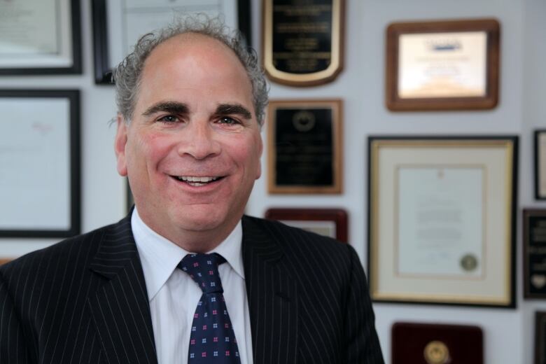 Man wearing a suit smiles, he is standing in front of a wall filled with various plaques. 