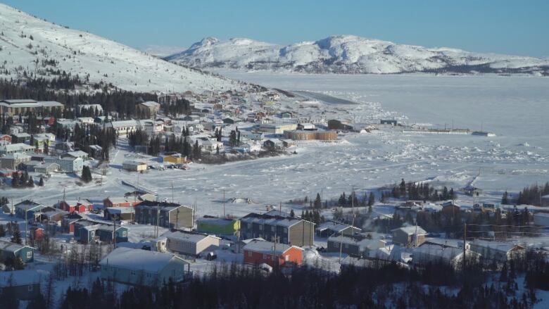 The houses of a small town, as seen by air, laid out in the foregroud, with snowy mountains and ocean in the background.