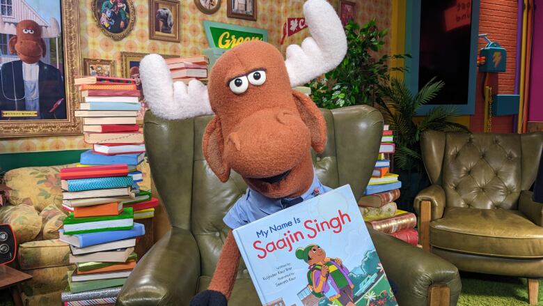 A moose puppet holds up a children's picture book, smiling.