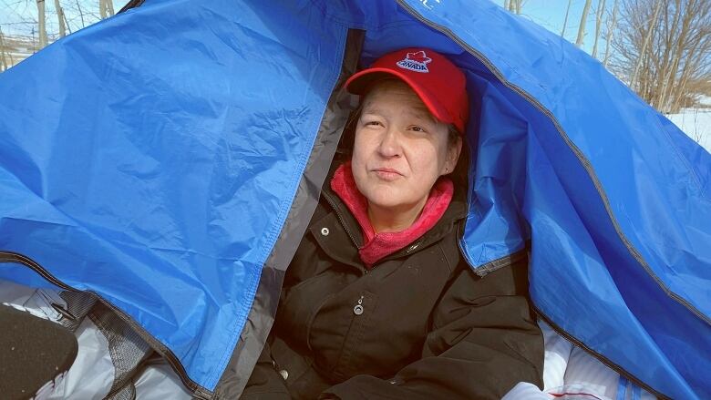 A woman with a red hat sits inside a blue tent in the snow and looks out.