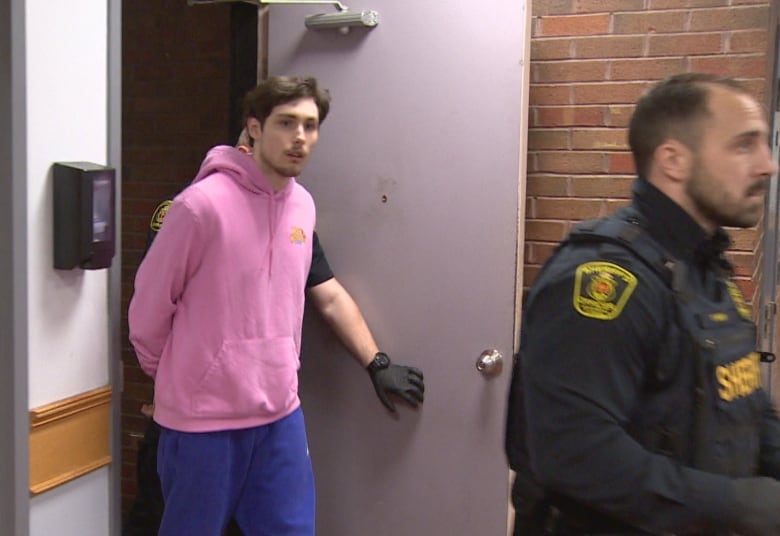 A man wearing a pink hoodie is guided in handcuffs by a sheriff.