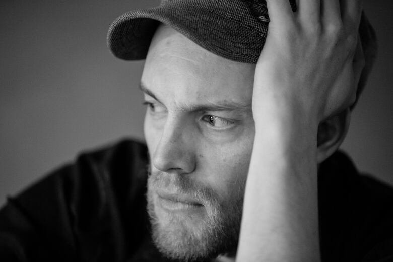 A pensive bearded man wearing a baseball cap holds his head up with his left arm.