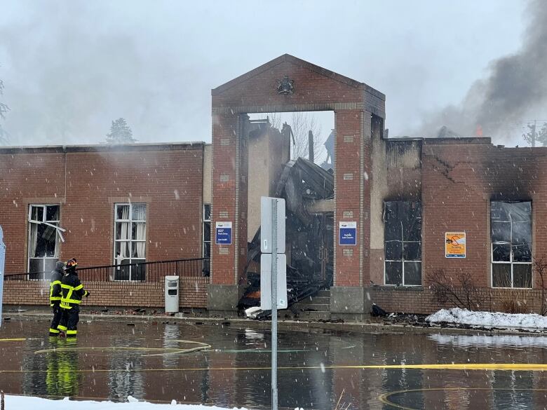 Two firefighters stand near a burned building with smoke coming out of it.