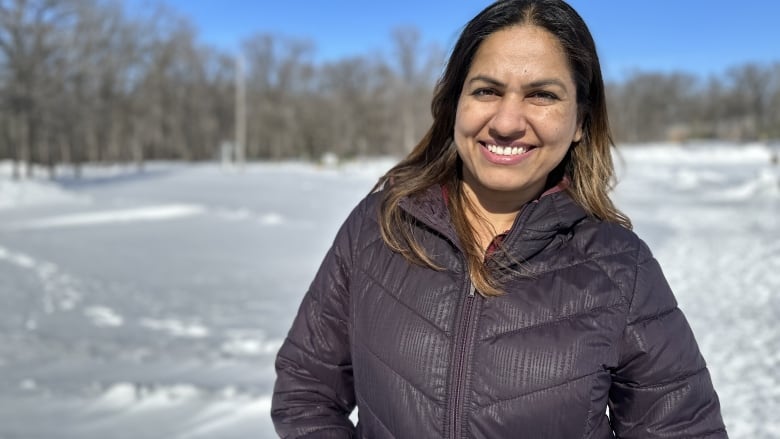 A woman in a black coat looks at the camera and smiles. She stands outside in a snowy park.