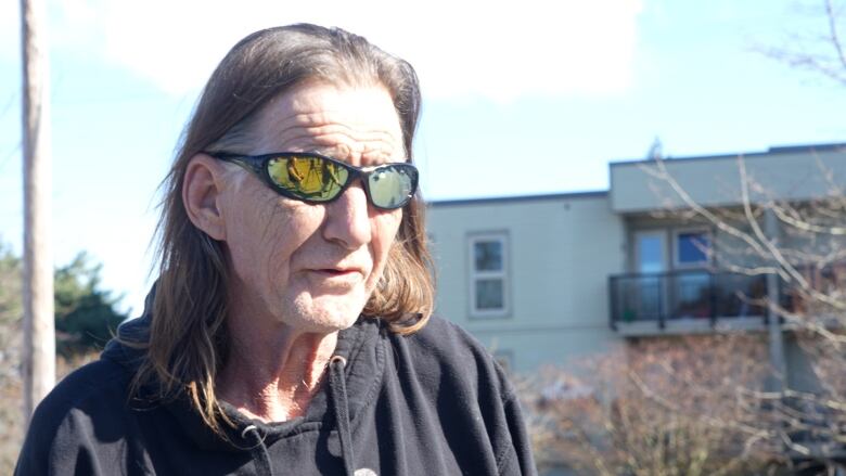 A man with sunglasses and long hair speaks to a reporter off-camera outside a building.