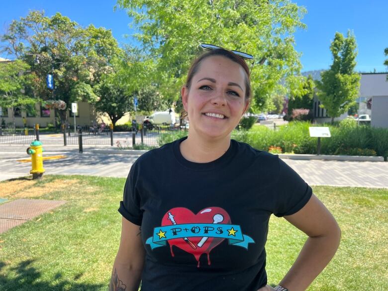 A woman in a black T-shirt with sunglasses on her head stands smiling in front of a grassy area with trees.