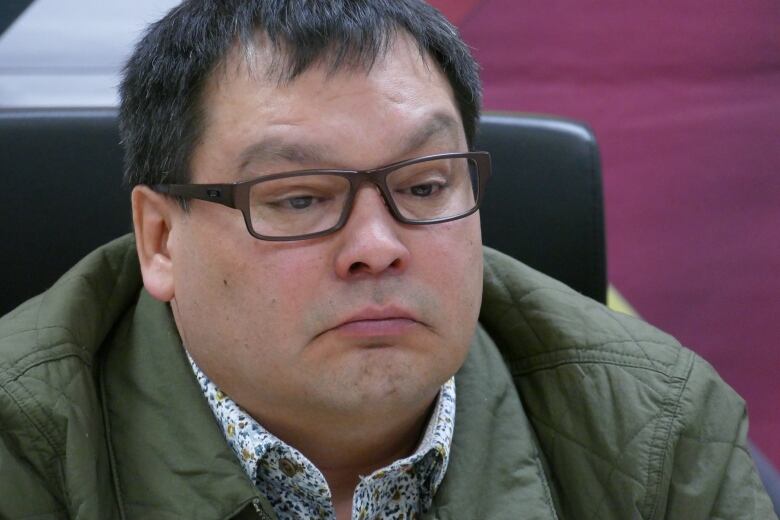 A man with short black hair, dark frame glasses and a green jacket speaks into microphones at a media event.