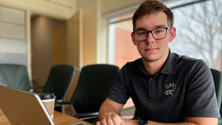 Adam MacKenzie, President of the UPEI Student Union, sits by his laptop in the union office.  He's wearing a grey, UPEI polo shirt. 