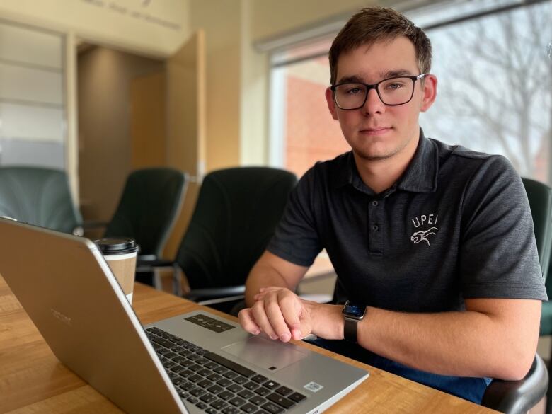 Adam MacKenzie, President of the UPEI Student Union, sits by his laptop in the union office.  He's wearing a grey, UPEI polo shirt. 