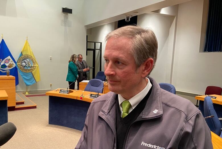 A mean wears a jacket while speaking into a microphone inside Fredericton council chambers.