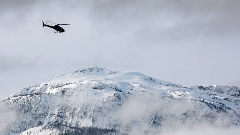 A helicopter flies under dark clouds over a snowy mountainside. 