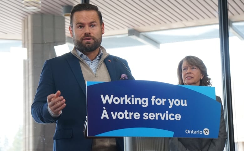 A man speaks into a microphone behind a sign that says, 