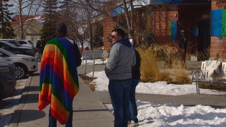 a group of three people stand outside a brick building. one is draped in a rainbow blanket