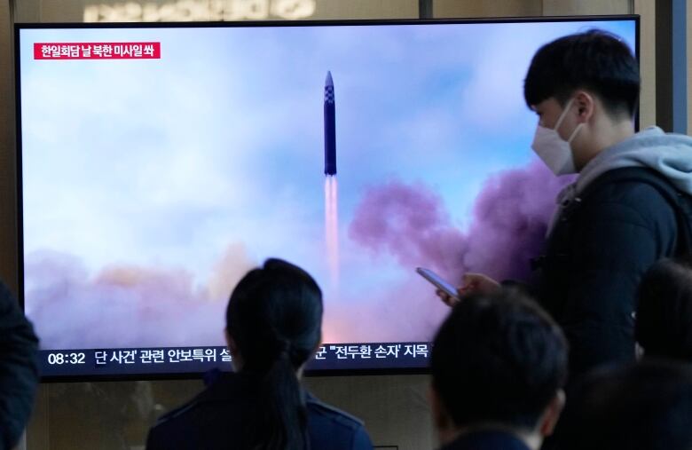 A television screen in a railway station shows an image of a missile being launched. Three passersby watch the screen. 