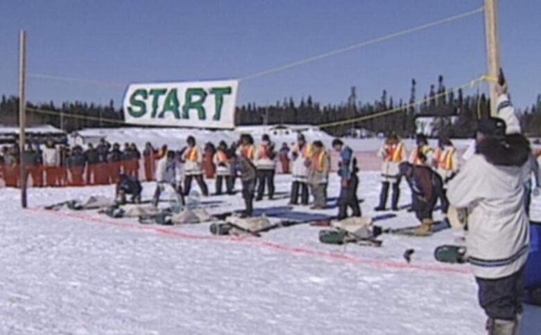 A group of racers stand on snow covered terrain at a starting line. 