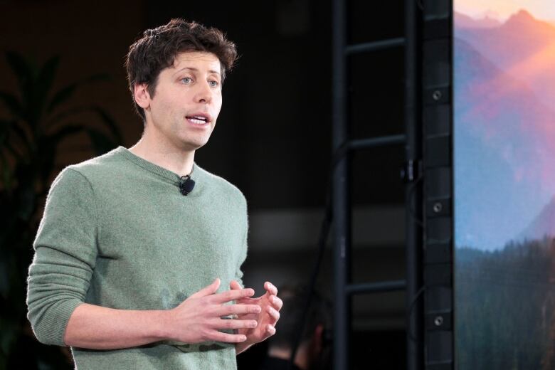 Man in green sweater gestures to offscreen audience while giving a presentation.