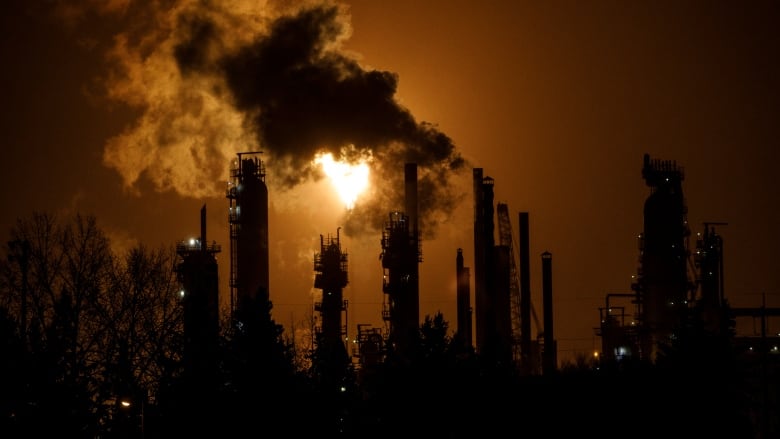 Smoke billows from smoke stacks against a dark sky. A bright orange flare lights up a portion of the sky.