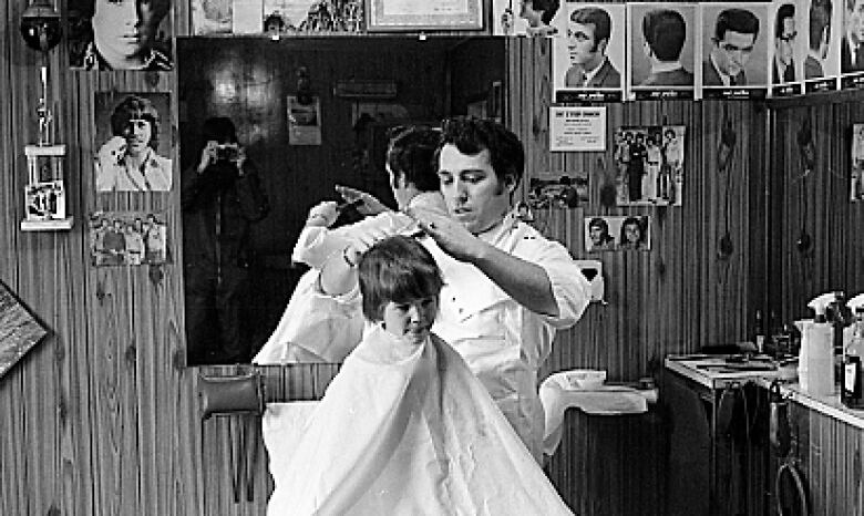 A black and white photo of a barber giving a haircut to a child. The photographer can be seen in the mirror holding the camera to his face.