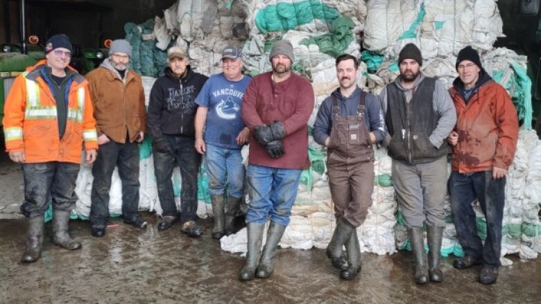 Eight men wearing rubber boots and other farmers' clothing stand in front of a large pile of bundled plastic