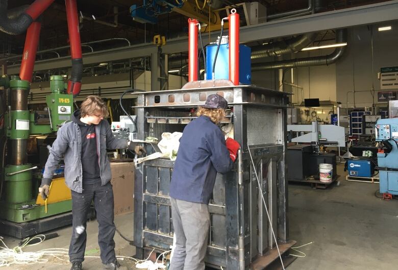 Two young people work in a large, industrial shop. The students are loading a large metal compactor with plastic.