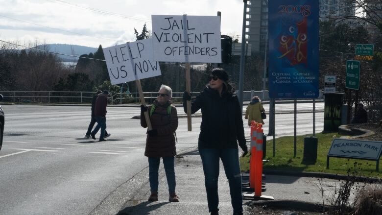 Two women are holding placards that read 'jail violent offenders' and 'help us now' on the sidewalk.