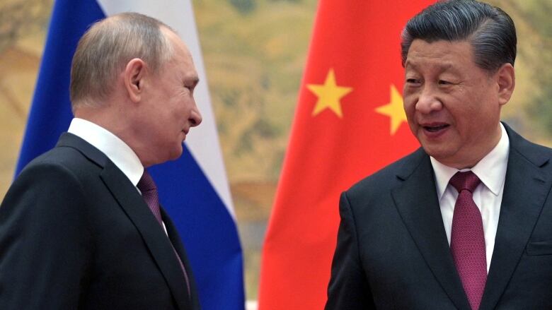 Two men wearing dark suits and purple ties smile at eachother as they stand in front of two flags.