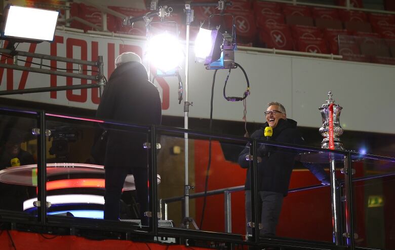 A man with white hair, holding a yellow microphone laughs as he stands on the set of an outdoor TV studio inside a soccer stadium. A trophy is shown next to him.