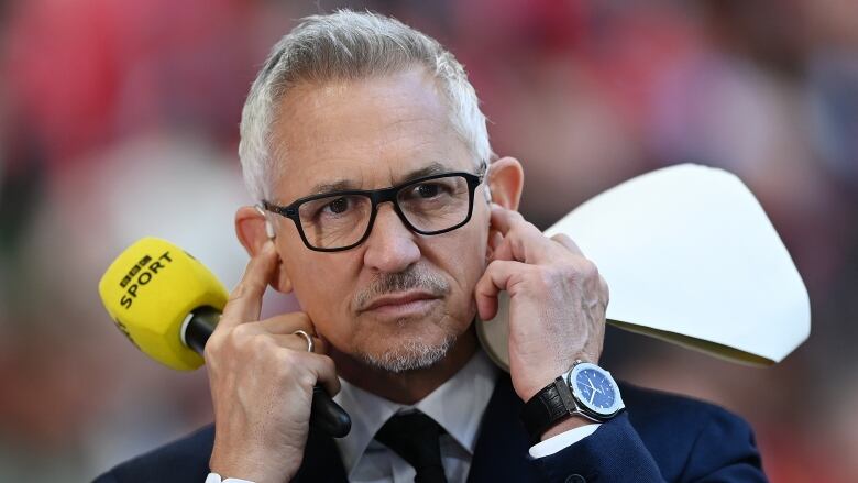 A man with white hair, wearing a suit and tie and black-rimmed glasses, plugs his ears with his fingers as he holds a microphone in one hand and a piece of paper in the other.