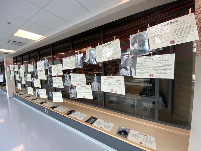 An exhibit featuring dozens of paper diplomas and photographs hang in a hallway at a university 