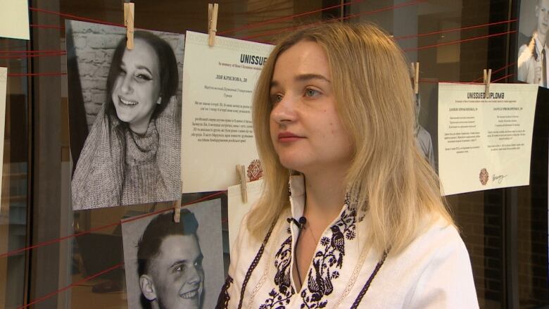 Yaryna has shoulder length blonde hair and is wearing a white shirt with brown and cream embroidery. She is standing next to a photograph of a student in the exhibit. 