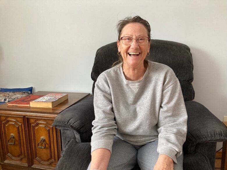 A woman with brown hair and a grey sweatshirt sits on a chair in a living room and laughs.