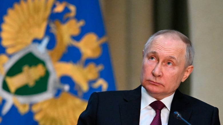 Wearing a dark suit, Russian President Vladimir Putin sits looking up at a blue flag. 