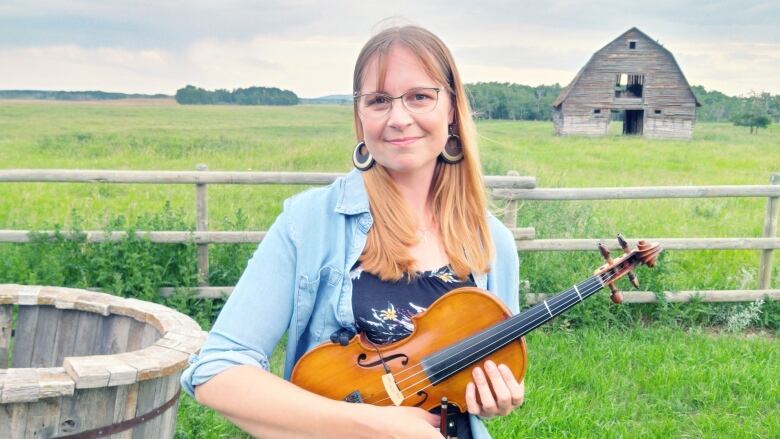 A woman holds a fiddle and stands in a rural scene with a barn behind her.