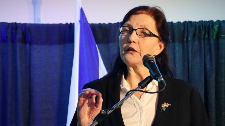 A woman in glasses speaks into a microphone and gestures with her hand.