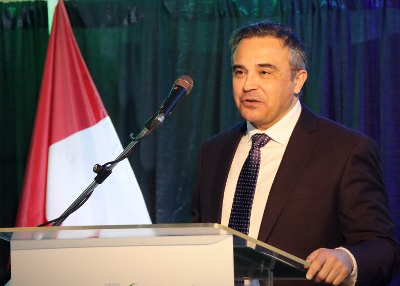 A man in a dark suit and tie smiles and speaks into a microphone.