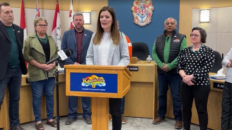 Seven people surround one woman speaking at a podium.