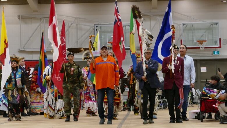 A crowd of people are walking toward the camera inside a gym, carrying flags, eagle staffs, some wearing ceremonial Indigenous and military clothing.