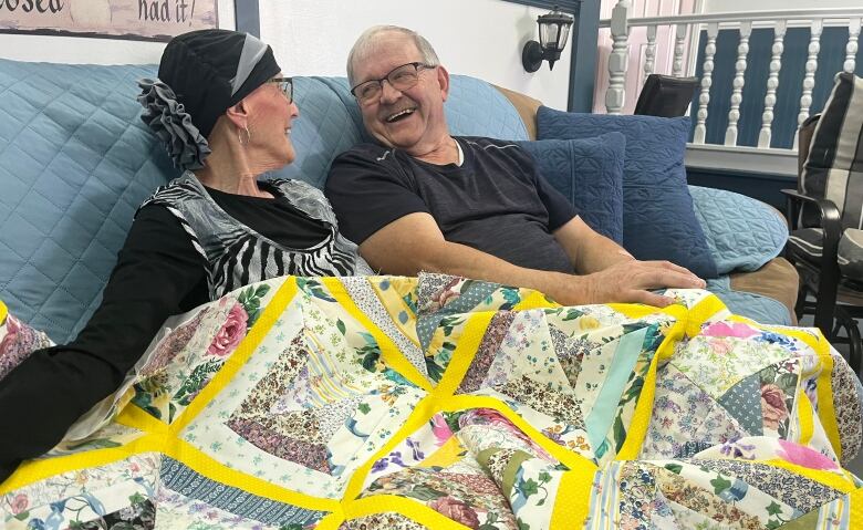 A man and a woman sit on a couch under a yellow quilt. 