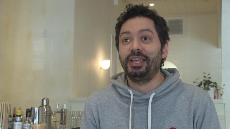 Bardia Illbeigi, a Persian man with a dark scruffy beard and short curly hair, is pictured from the shoulders up, wearing a grey hoodie and standing in his restaurant. 