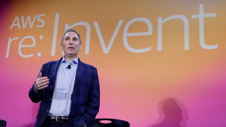 A man is shown on a stage speaking while standing in front of a backdrop with a logo.