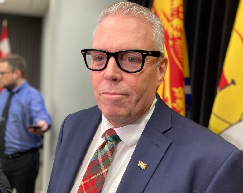 A man with grey hair and glasses wears a blue suit and a tartan tie. 