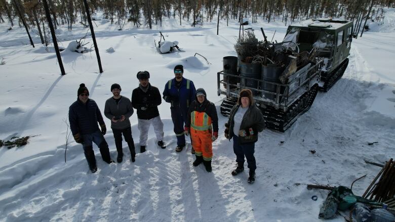 An aerial photo of a group of men standing in front of a tracked vehicle carrying a trailer worth of burned material. The landscape is wintery and forested.