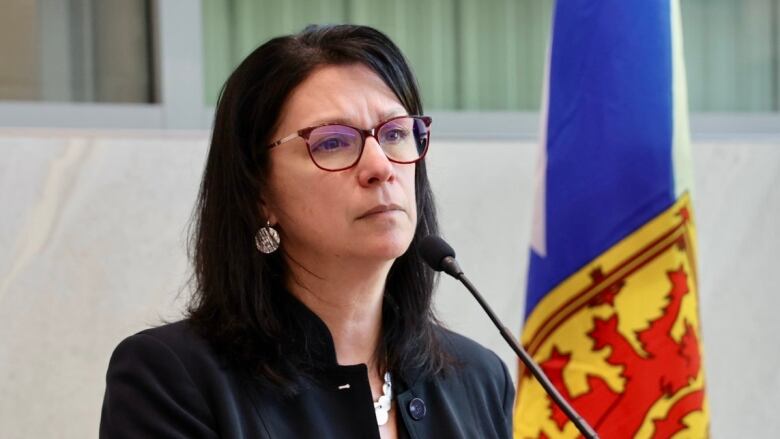 A woman with shoulder-length dark brown hair and glasses and silver earrings and a silver necklace wears a black jacket. She is standing next to a Nova Scotia flag. 